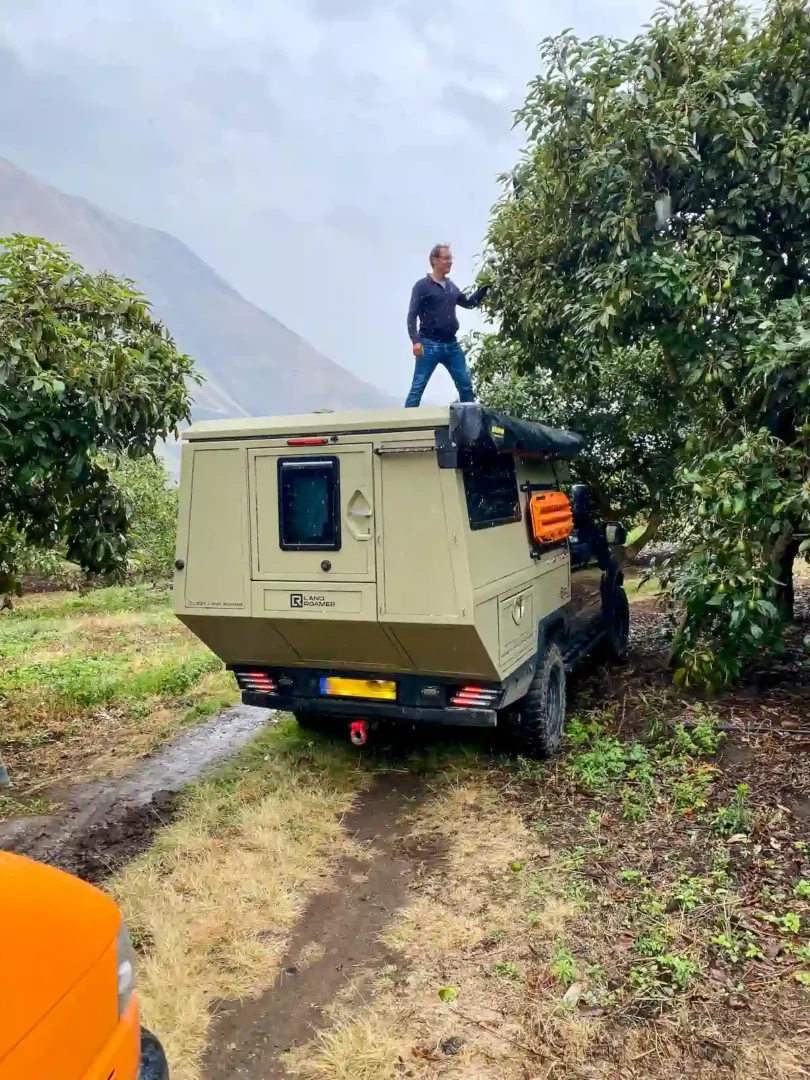 Una persona subida en la célula vivienda Pioneer de Land Roamer, apartando unas ramas para poder pasar con su pickup overland